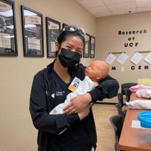 Kaitlyn Yu, a junior nursing student at UCF, wearing black College of Nursing scrubs holding a simulation newborn manikin in a classroom.