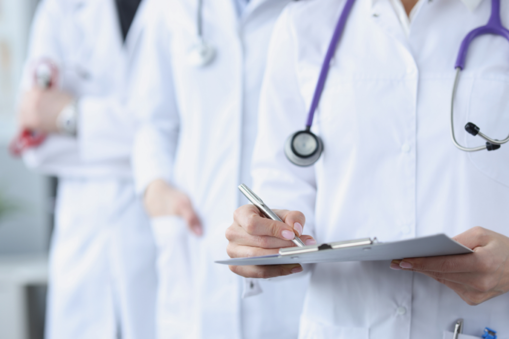 Three people in white clinical coats with stethoscopes and a clipboard (no faces seen)