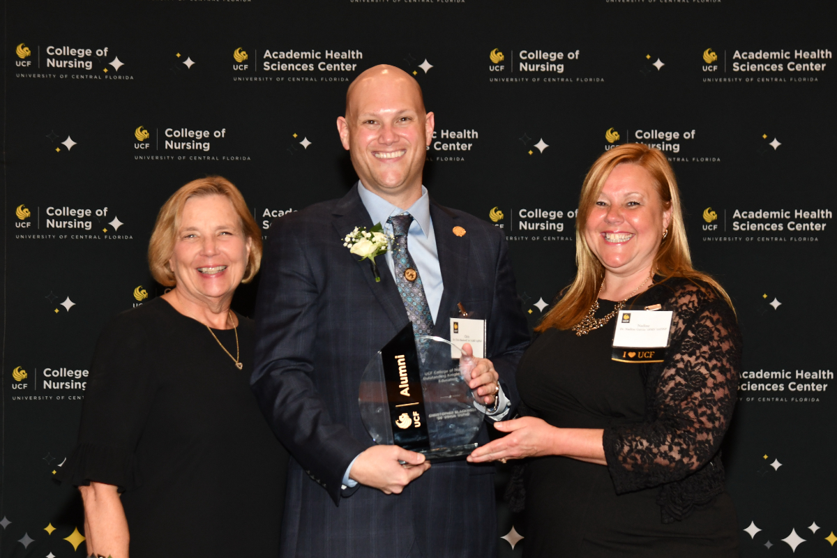 2023 Outstanding Knight Nurse Awardee Christopher Blackwell holds a glass award and stands between UCF College of Nursing Dean Mary Lou Sole and UCF College of Nursing Alumni Chapter board member Nadine Garcia.