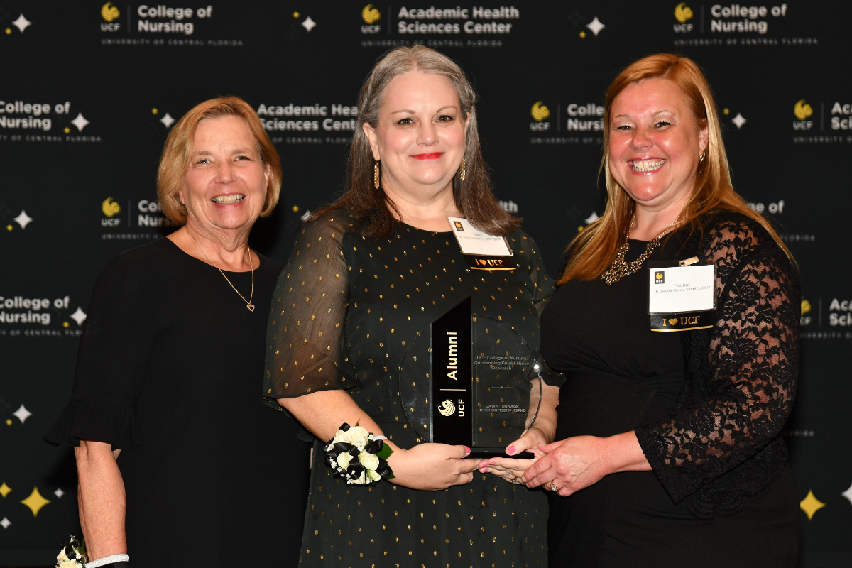 2023 Outstanding Knight Nurse Awardee Dawn Turnage holds a glass award and stands between UCF College of Nursing Dean Mary Lou Sole and UCF College of Nursing Alumni Chapter board member Nadine Garcia.