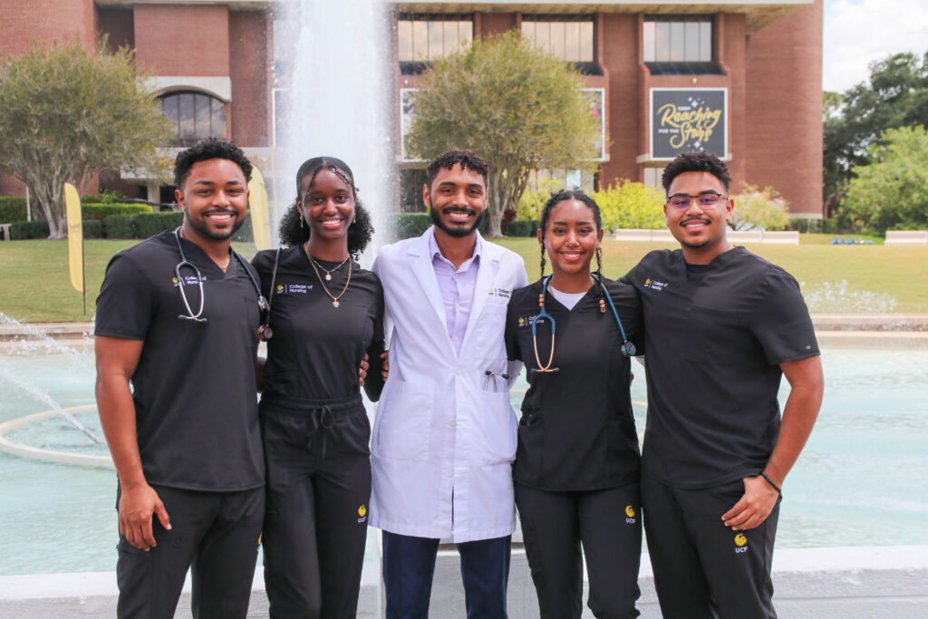 4 UCF Nursing students in black clinical scrubs and one student in the center wearing a white clinical lab coat stand in front of the UCF reflection pond. The five students are cousins.