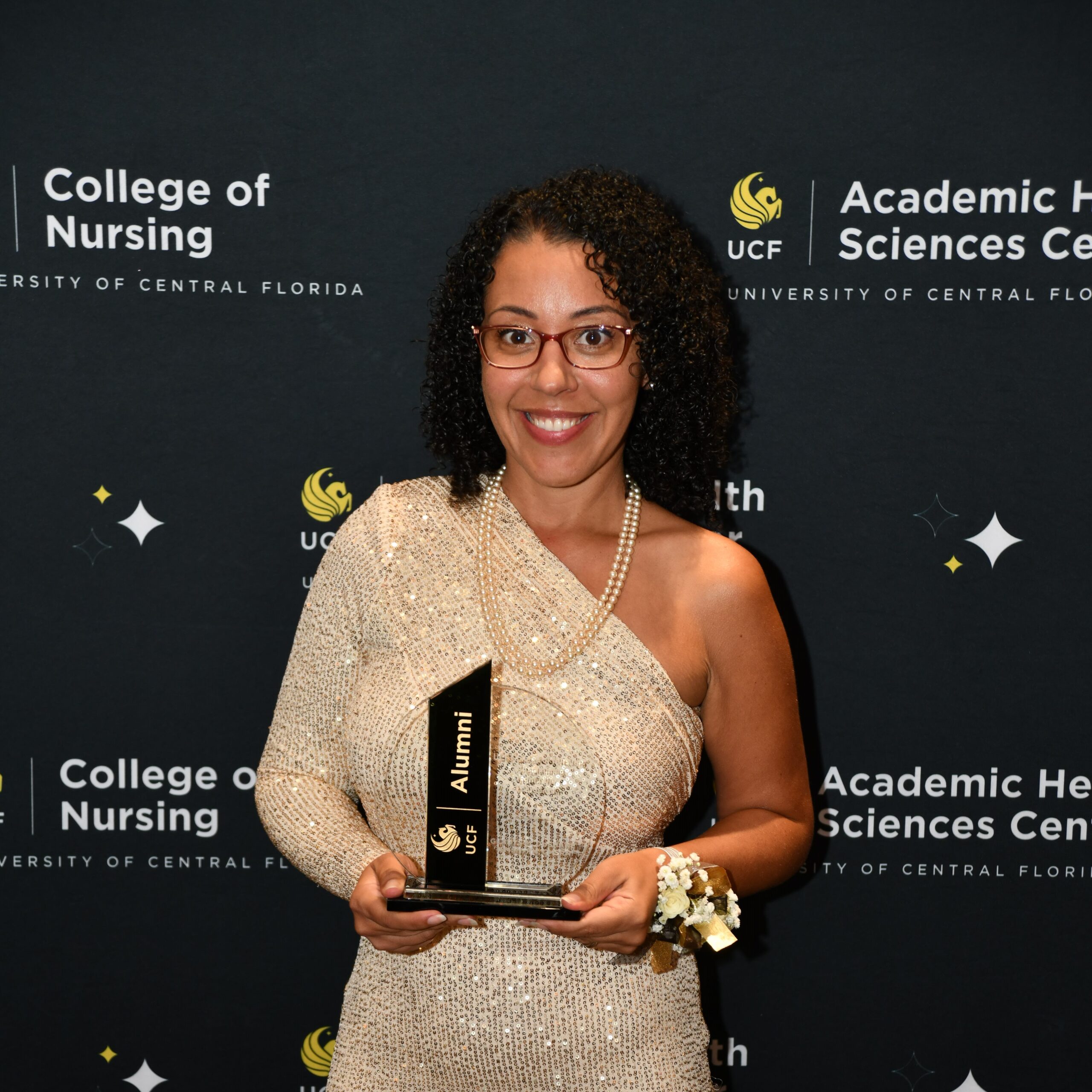 Gladys Dushane holding a crystal UCF Alumni award in front of a UCF College of Nursing banner
