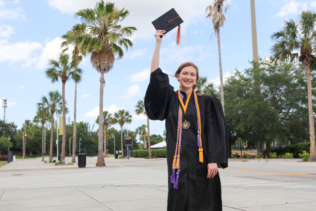 Rebekah May stands in regalia holding a graduation cap in the air on a sidewalk in front of rows of palm trees on UCF's Orlando campus.