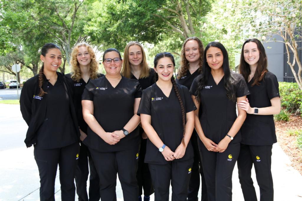 Eight female UCF nursing students in black logo scrubs stand outside with trees in the background.