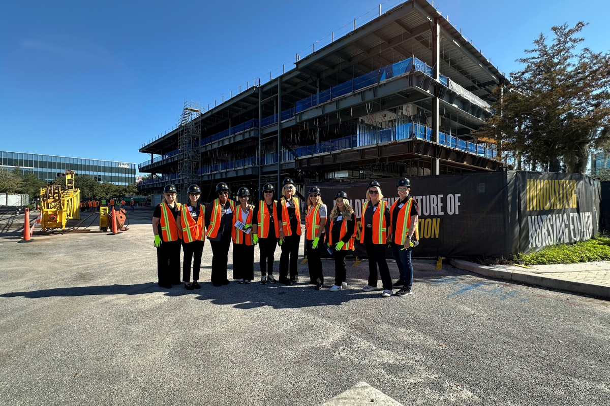 A group from Orlando Health wearing construction vests, gloves and hard hats stands in front of the Dr. Phillips Nursing Pavilion, which is under construction in Lake Nona on a clear blue sky day in Florida.