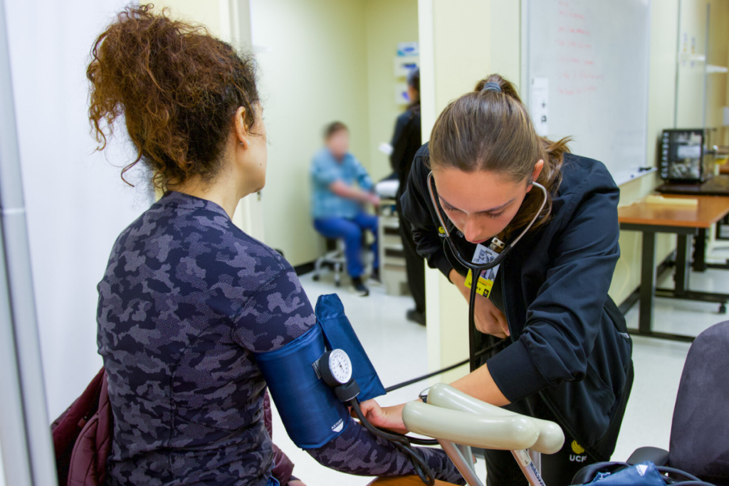 A UCF nursing student in black clinical scrubs checks blood pressure on an embedded participant during a rural health clinic simulation at UCF's STIM Center