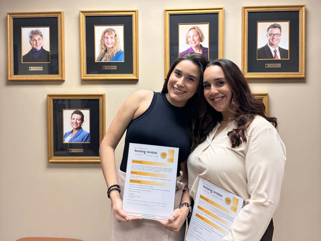 Two students holding Nursing Unidos flyers in front of an endowed faculty photo wall at UCF College of Nursing