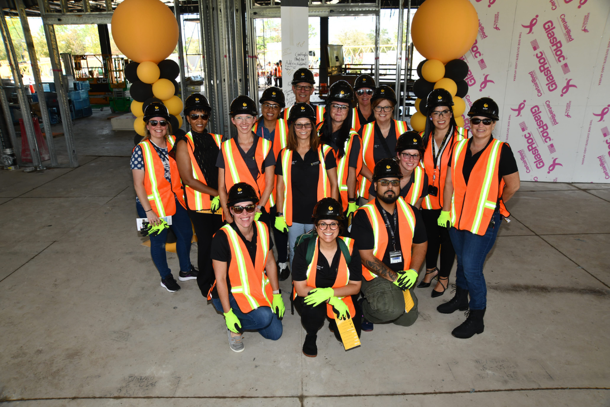 A group of UCF faculty and staff wearing construction vests, gloves and hard hats pose in front of balloons and a steel column inside the Dr. Phillips Nursing Pavilion, which is under construction in Orlando, Florida