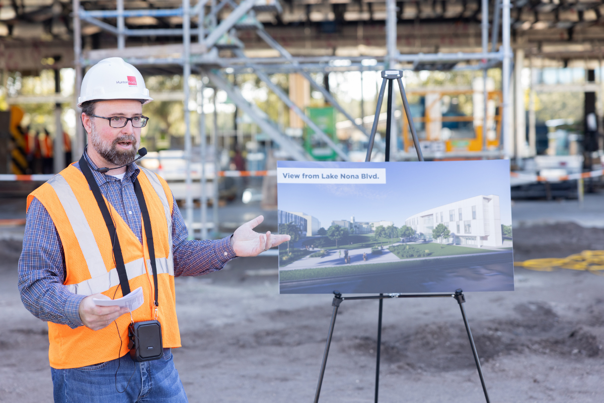 A member from the construction team speaks in front of the Dr. Phillips Nursing Pavilion, which is under construction, and next to an easel of the building rendering