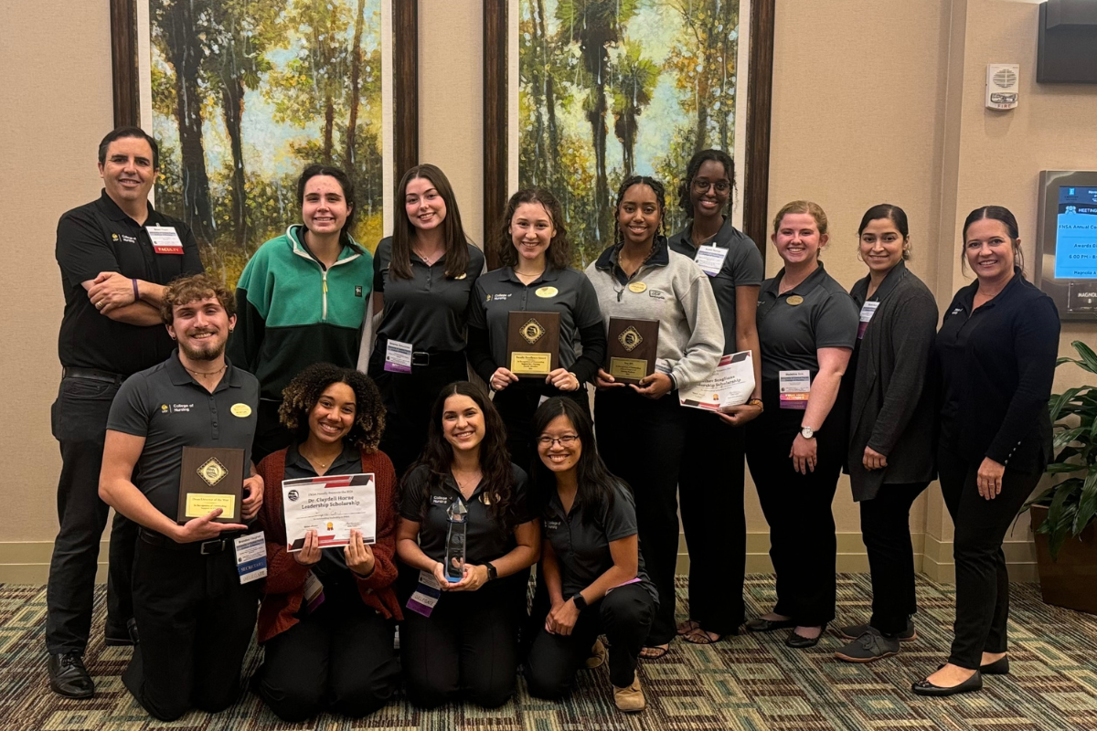 A group of UCF College of Nursing students wearing school polos hold certificates and stand with two faculty advisors inside a hotel ballroom.