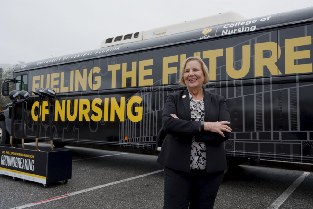 Dean Mary Lou Sole stands with arms folded smiling in front of a UCF bus with Fueling the Future of Nursing graphics at the Dr. Phillips Nursing Pavilion groundbreaking event.