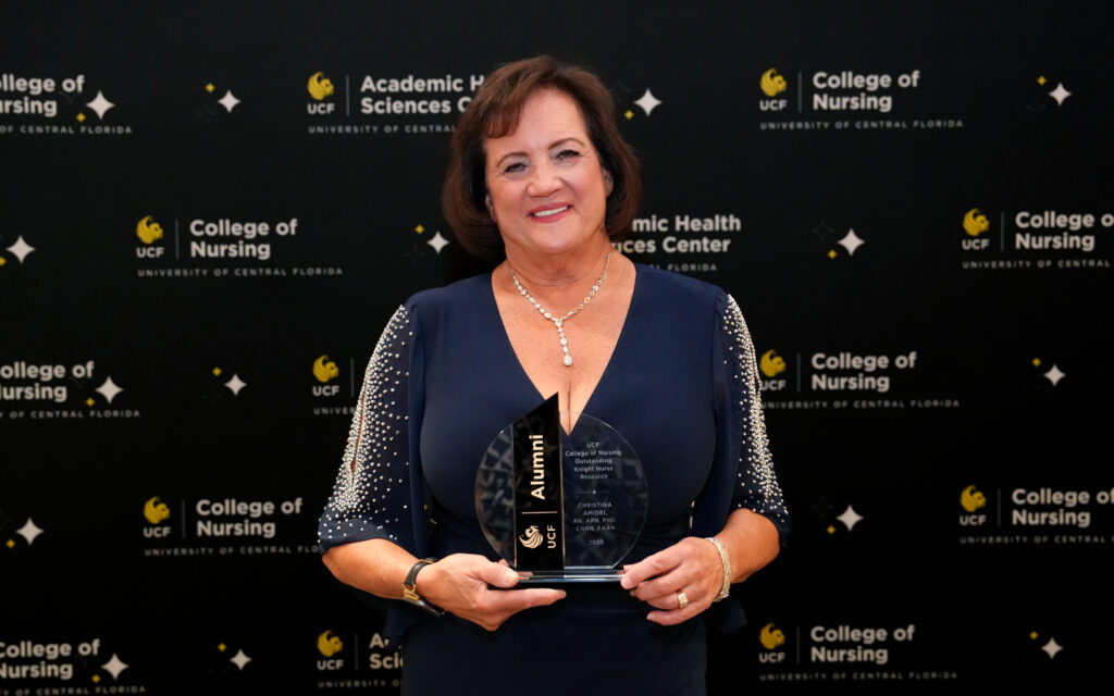 Christine Amidei holding a crystal UCF Alumni award in front of a UCF College of Nursing banner