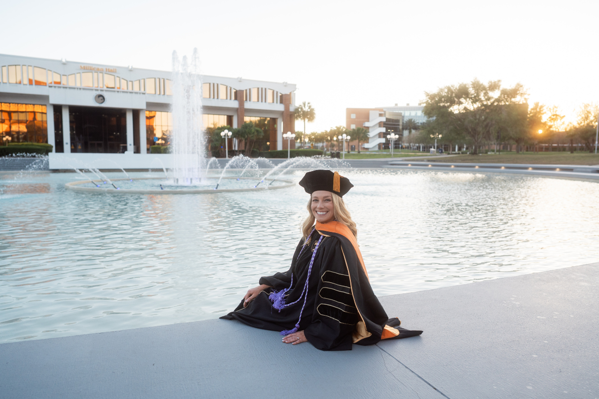 Elizabeth Kattleman wears UCF regalia and smiles in front of the reflection pond at Millican Hall on campus in Orlando, Florida.