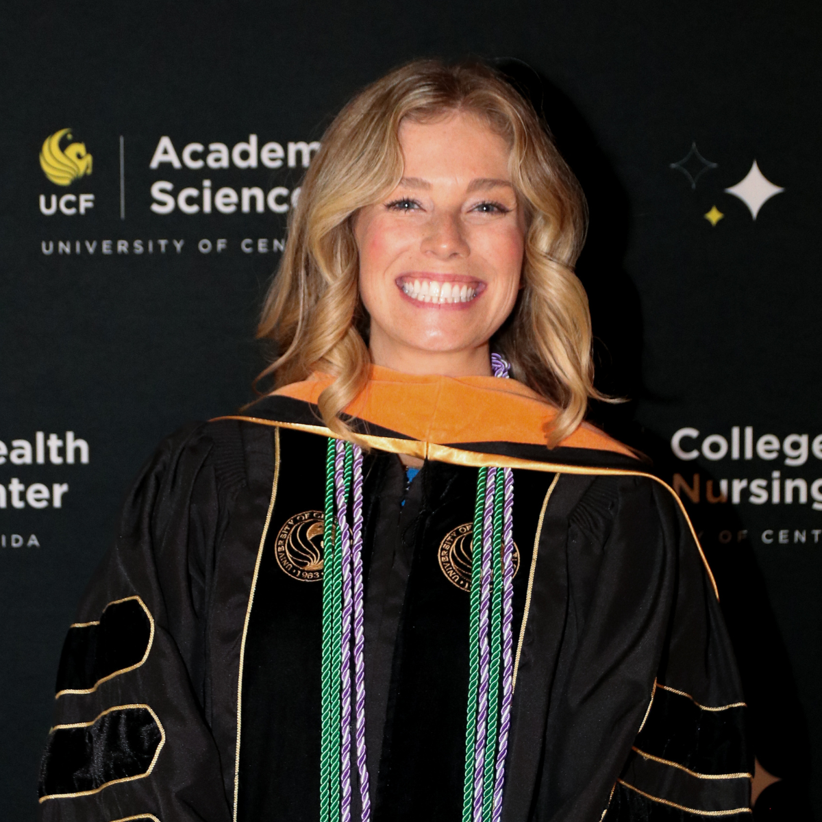 Elizabeth Kattleman, a Spring 2025 UCF doctor of nursing practice graduate, smiles in regalia in front of a UCF pegasus banner.