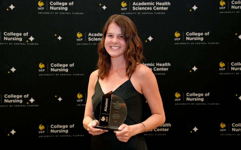 Kristine Gibbons holding a crystal UCF Alumni award in front of a UCF College of Nursing banner