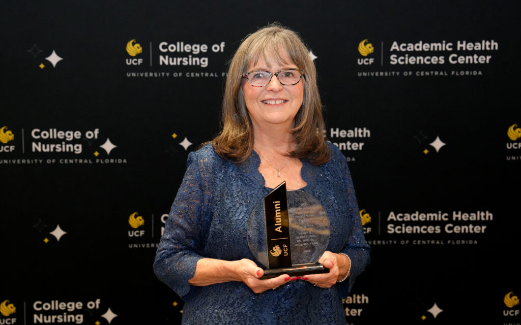 Mary G. Harper holding a crystal UCF Alumni award in front of a UCF College of Nursing banner