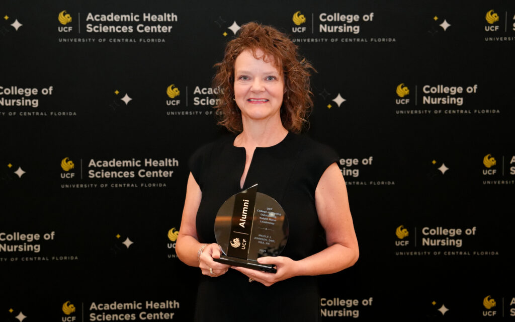 Nicole J. Johnson holding a crystal UCF Alumni award in front of a UCF College of Nursing banner