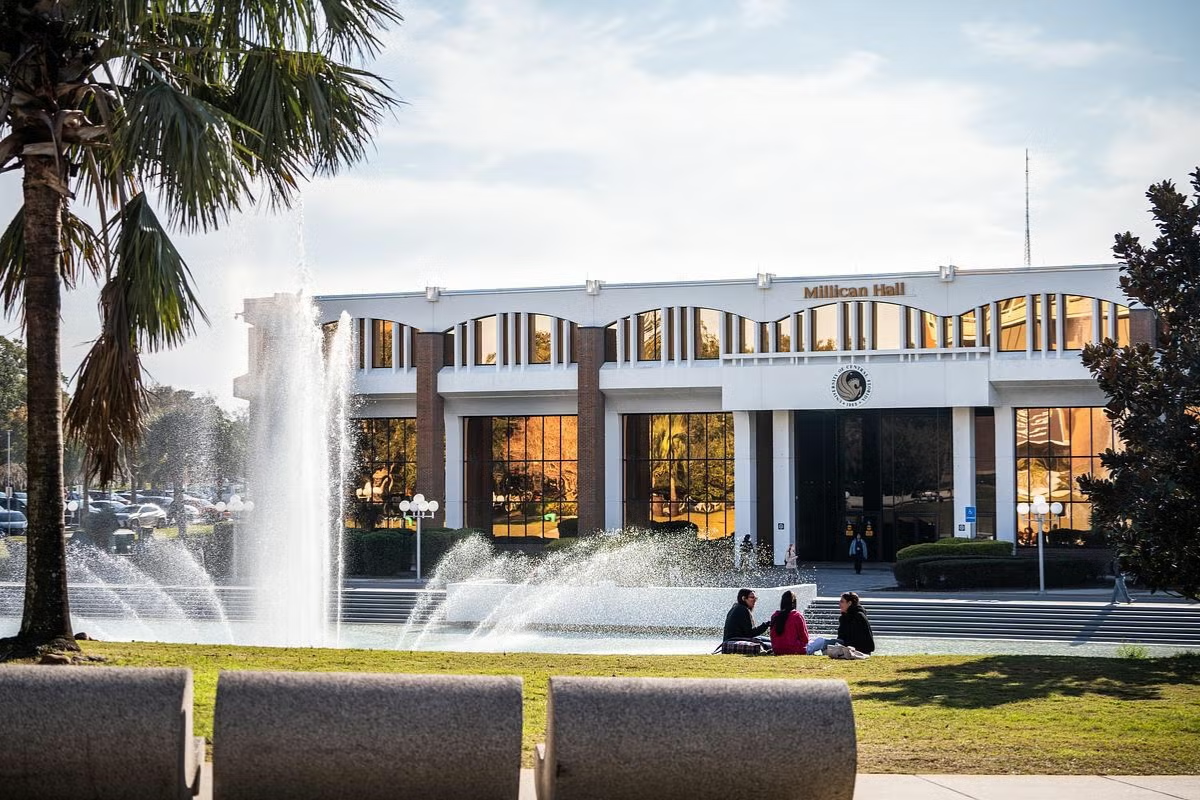 Three students sit on the lawn in front of the reflection pond facing Millican Hall on UCF's main campus in Orlando, Florida.
