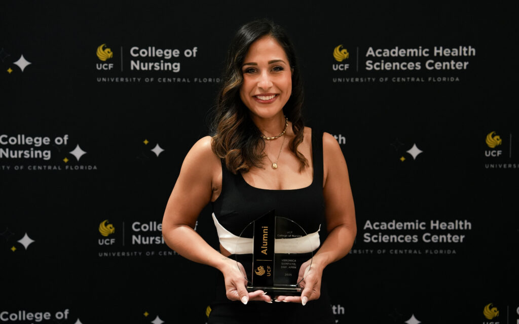 Veronica Sampayo holding a crystal UCF Alumni award in front of a UCF College of Nursing banner