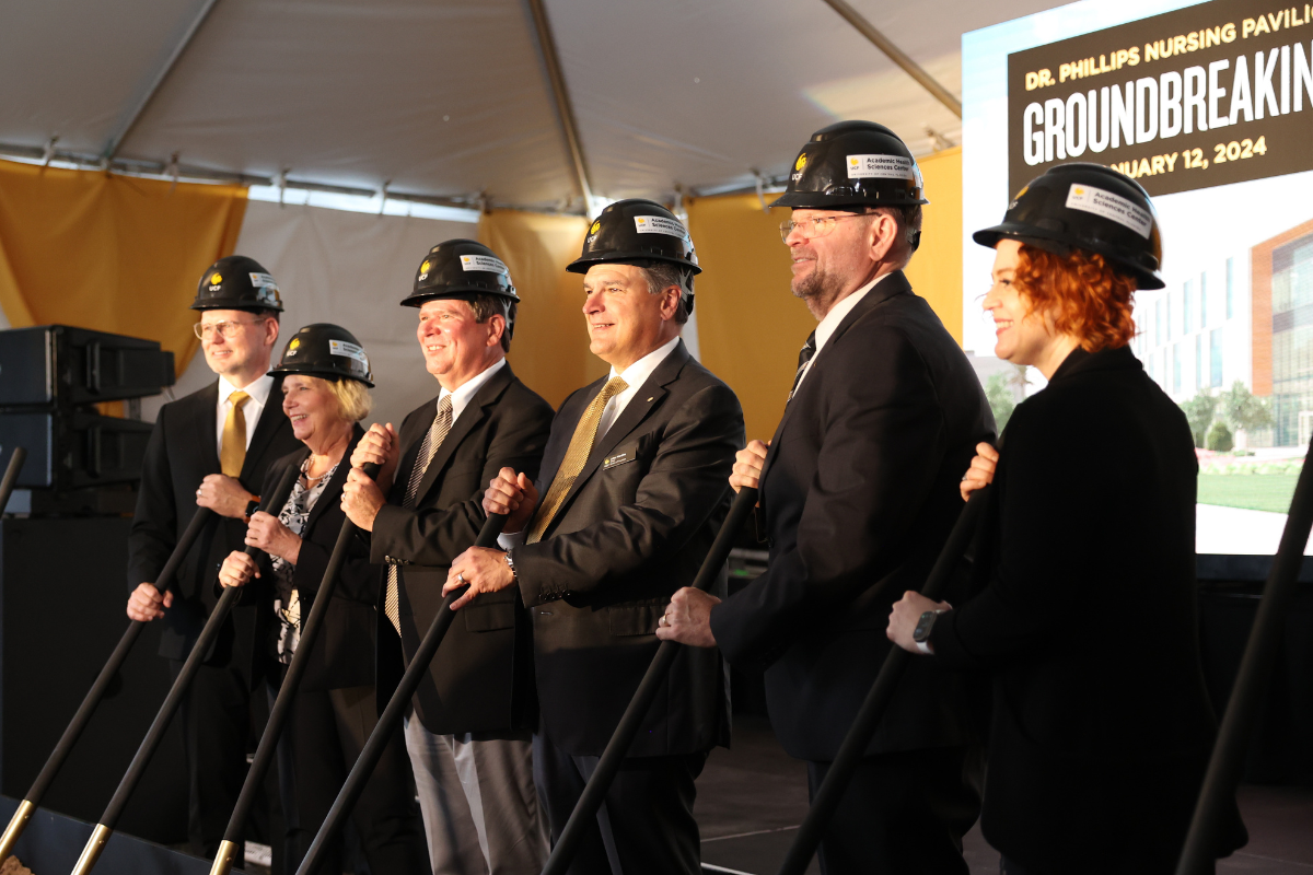 Six men and women in black business suit wearing UCF hardhats smile holding shovels at the Dr. Phillips Nursing Pavilion groundbreaking ceremony.