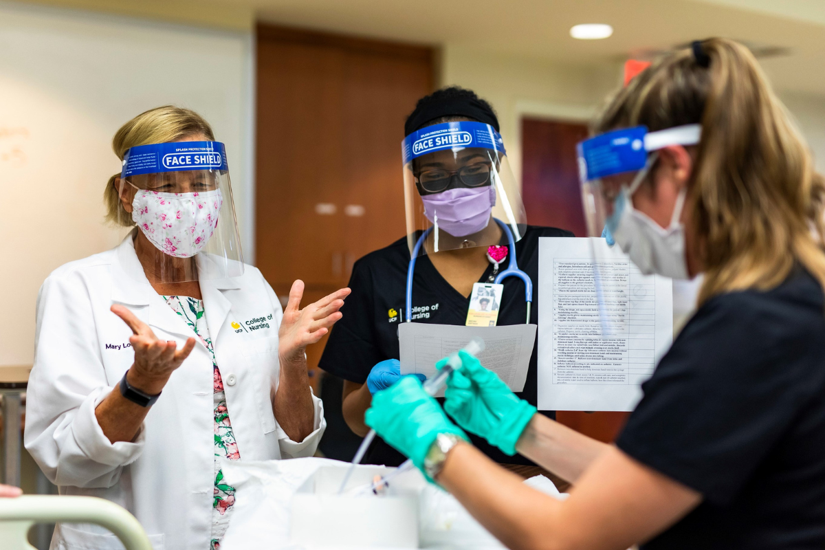 Dean Mary Lou Sole observes UCF nursing students in a simulation skills lab. All are wearing face masks and shields.