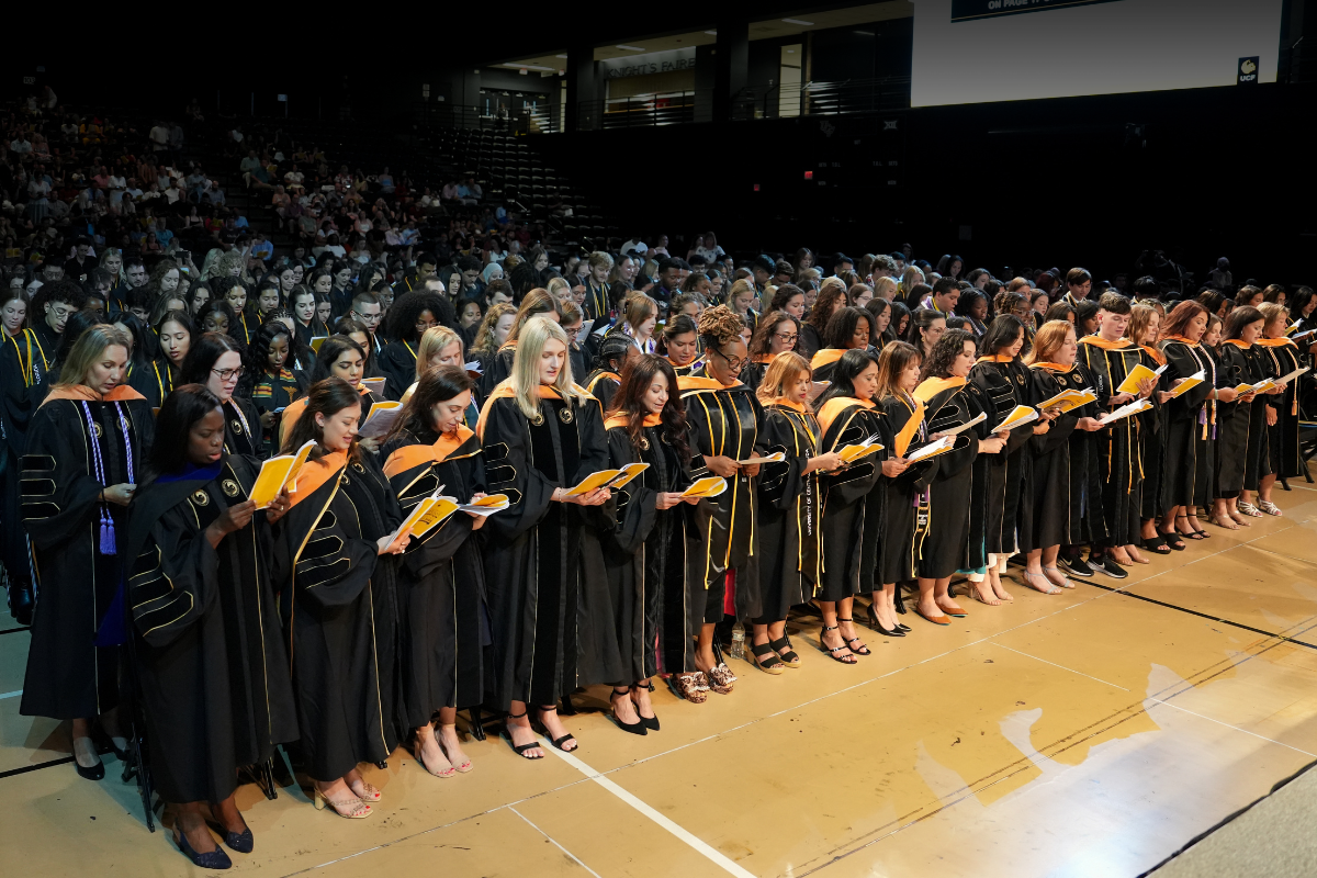 A group of students in regalia stand together and read from a program inside The Venue at UCF during Summer 2025 graduation celebrations.