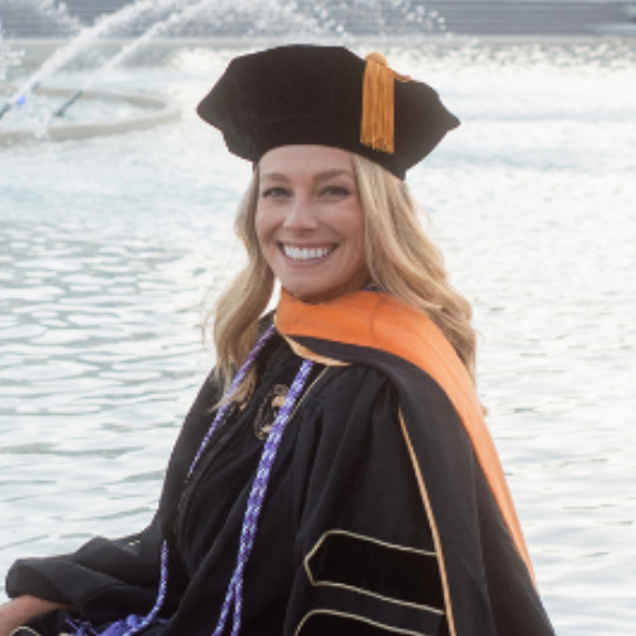 Elizabeth Kattleman wears UCF regalia and smiles in front of the reflection pond at Millican Hall on campus in Orlando, Florida.