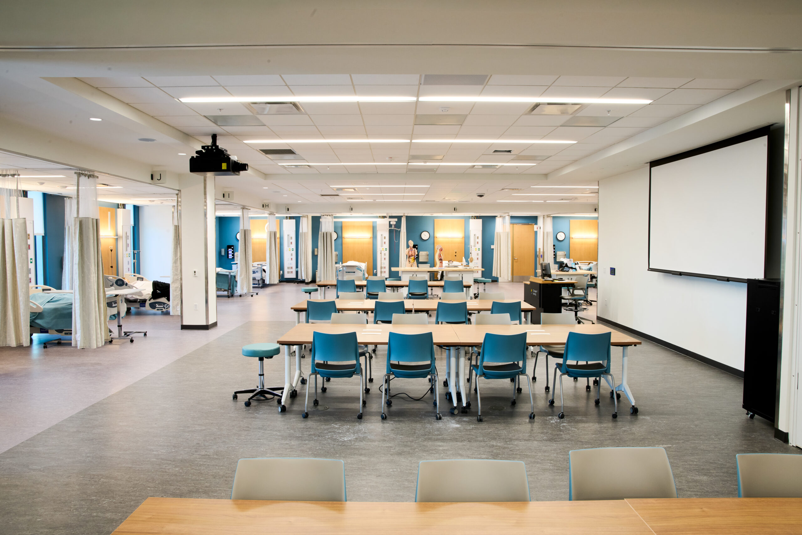 The Essentials and Skills Lab room showing simulated hospital rooms and a set of tables and chairs in the middle, along with a projection screen.