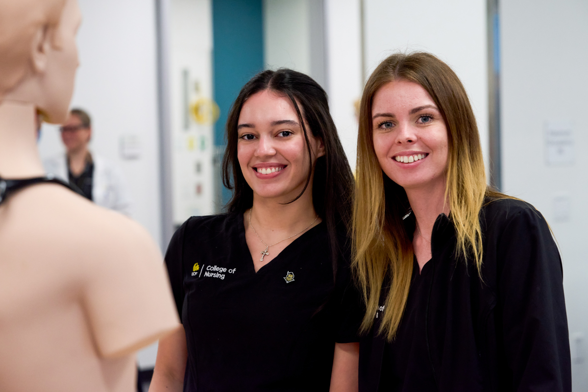 Two UCF College of Nursing students in black scrubs smile inside a simulation classroom